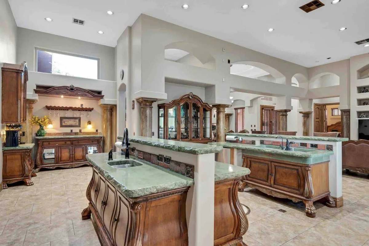 This image features a spacious chef’s kitchen with dual islands, decorative cabinetry, expansive granite countertops, and large windows that flood the space with natural light. The kitchen has an upscale, sophisticated feel and is arranged for both cooking and entertaining.