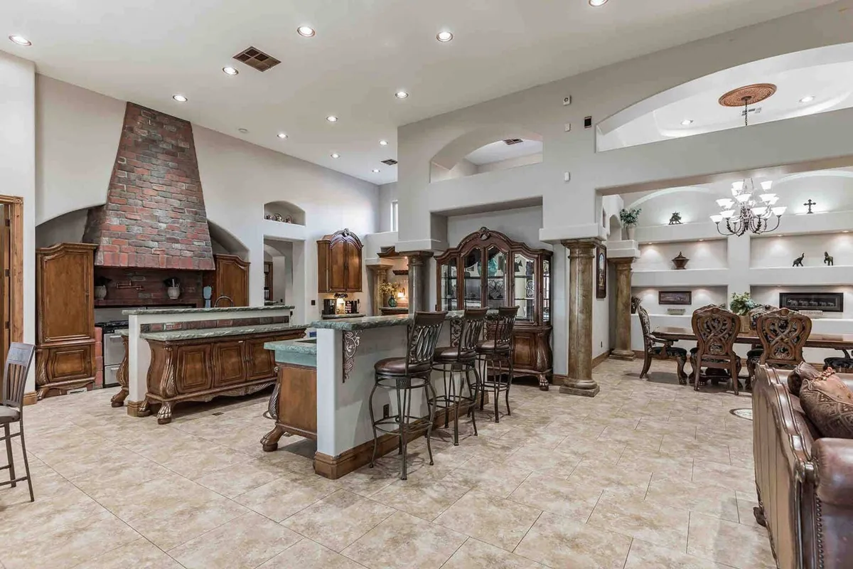 A grand open-concept interior highlights a large stone fireplace, kitchen island with bar seating, and an adjoining dining area set beneath arched doorways. The layout encourages both cooking and gathering, with the kitchen seamlessly transitioning into the elegant dining space.