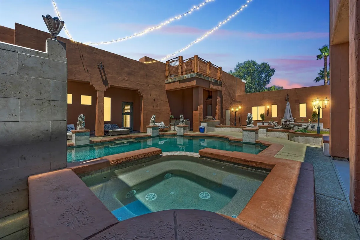 The image showcases the exterior of a luxurious single-family house in Glendale, Arizona, styled to resemble a castle. The focal points include a sparkling swimming pool and a bubbling hot tub, both elegantly framed by decorative statues and enhanced by warm outdoor lighting. A balcony overlooks the pool, inviting guests to relax and take in the vibrant sunset views. The overall mood is tranquil yet celebratory, perfect for evening gatherings, barbecues, and stargazing under the Arizona sky.