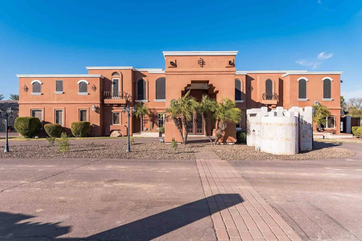 This exterior view showcases the main entrance with ornate architectural details, decorative windows, palm trees, and a striking stone structure. The scene conveys vibrancy and readiness for social gatherings or festive departures to nearby events.