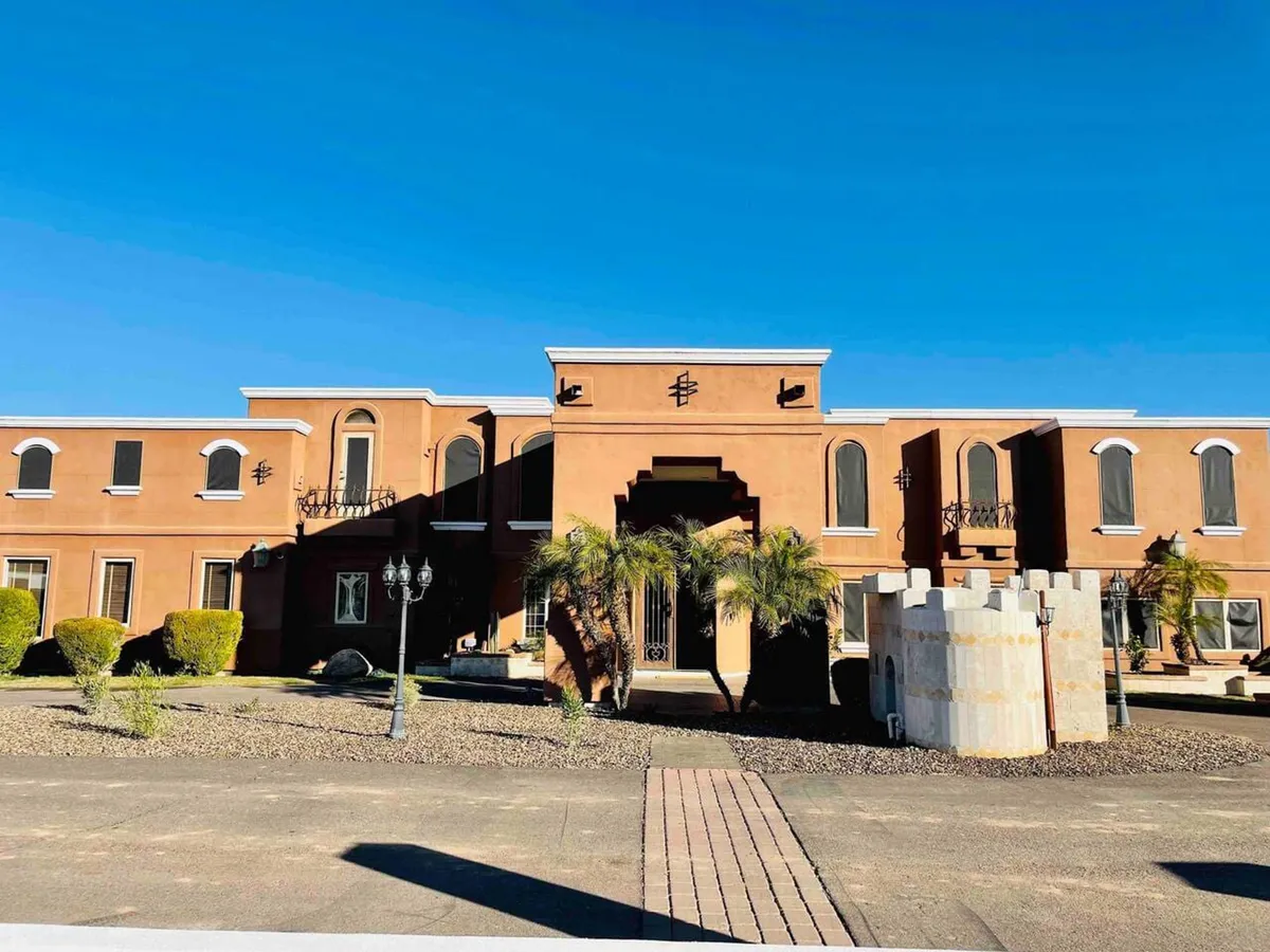 The image captures the home's impressive main entrance, framed by a decorative archway, palm trees, and a castle-like turret. Streetlamps and manicured landscaping add to the sense of arrival and grandeur, suggesting a gathering spot for groups before heading out to nearby events.