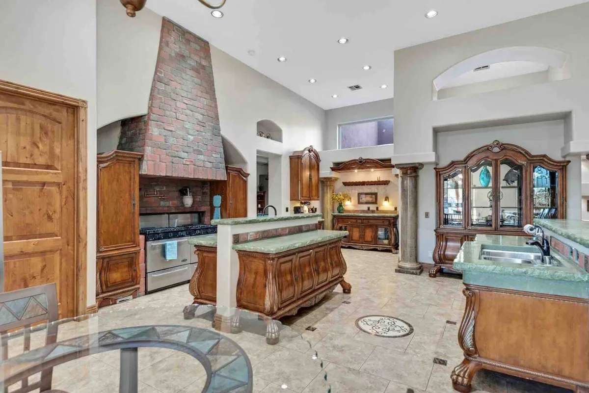 An inviting kitchen and dining area featuring a central island with a green countertop, large wooden cabinets, a brick chimney, and a spacious table. The arrangement suggests communal meals, pre-game celebrations, or post-event feasts.