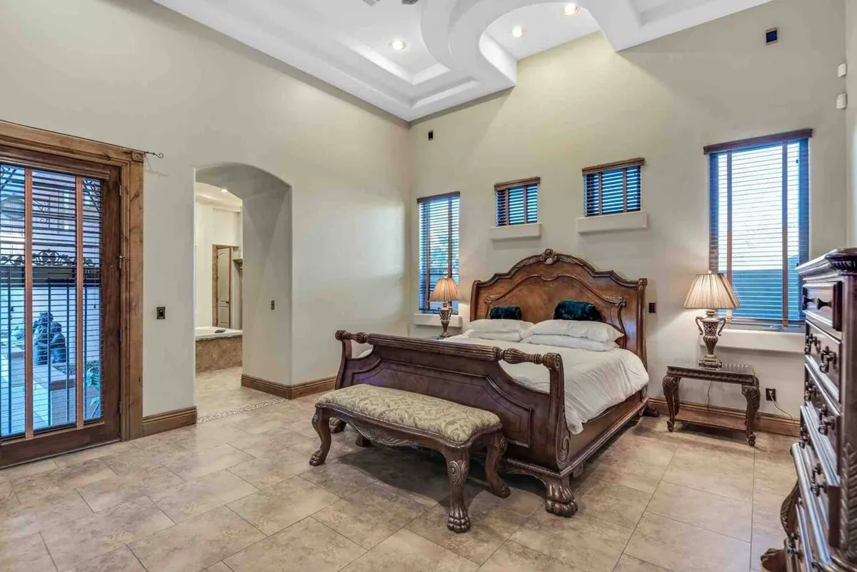 A serene and luxurious bedroom featuring an ornate wooden bed frame, bedside tables with lamps, decorative wall texture, and large windows with wooden blinds that let in natural light. The room exudes comfort and privacy, ideal for restful nights and morning relaxation.