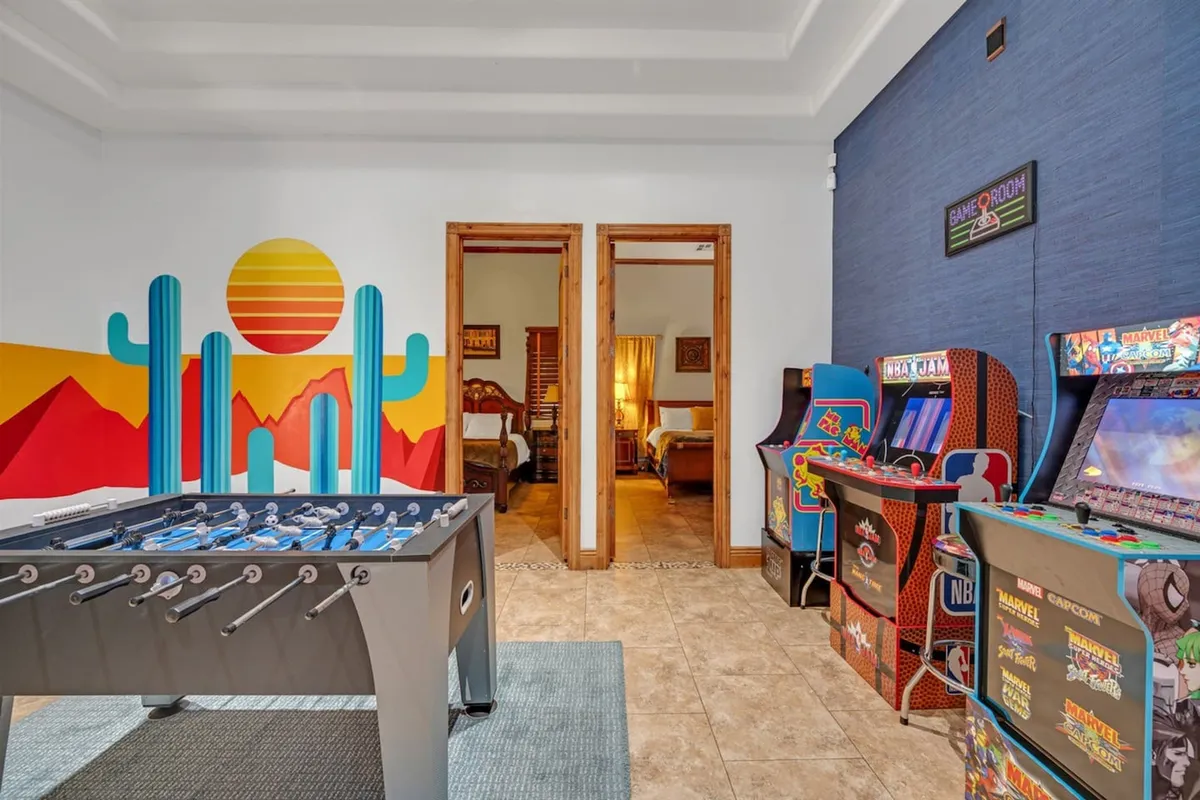 A vibrant arcade and game room featuring a foosball table, multiple arcade machines, a colorful wall mural, and visible doorways to other rooms, suggesting open flow and easy access between spaces.