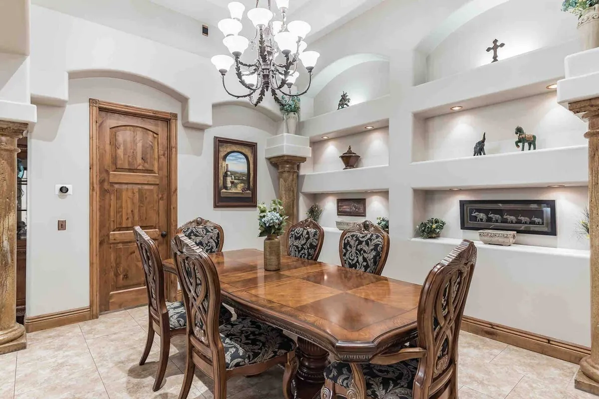 An elegant formal dining room with a wooden table, ornate dining chairs, a sparkling chandelier, and decorative wall niches. The space is set for a family gathering or festive celebration, exuding sophistication and warmth.